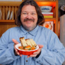 Matty Matheson holding an egg bake recipe topped with tomato sauce, potatoes, and sausage, served with cheesy bread.